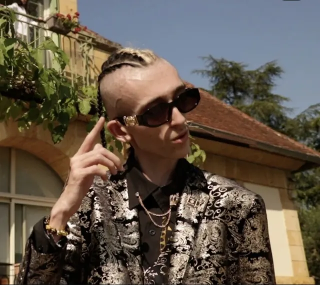 STONAN en veste baroque noir et or, lunettes de soleil, tresses blondes